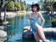 A woman in a blue dress and hat sitting on a rock in the water.